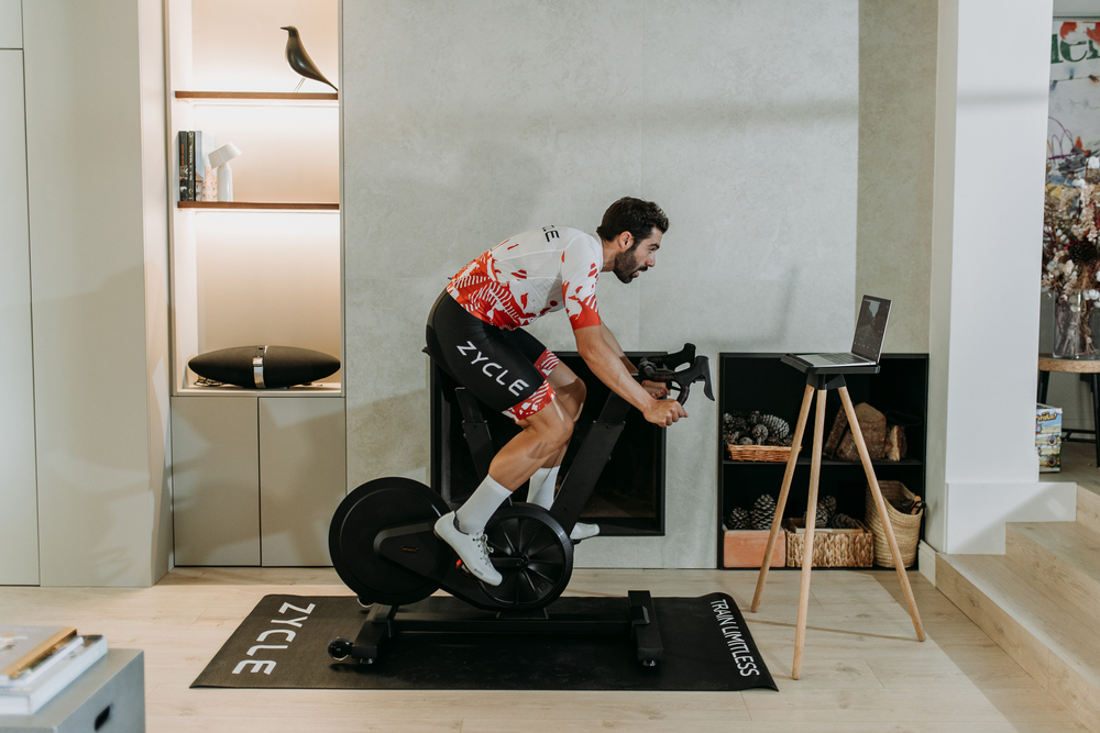 Ciclista sobre una bicicleta indoor conectada en un salón, con un portátil junto al equipo.